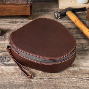 Op de afbeelding: Een bruine leren reisetui met een afgeronde vorm en een ritssluiting. De etui heeft een polsbandje en staat op een houten oppervlak. De ritssluiting heeft een donker bronzen kleur.