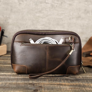 May include: A brown leather clutch with a zipper closure and a wrist strap. The clutch has a front pocket with a zipper and contains white charging cables. The leather has a distressed appearance.