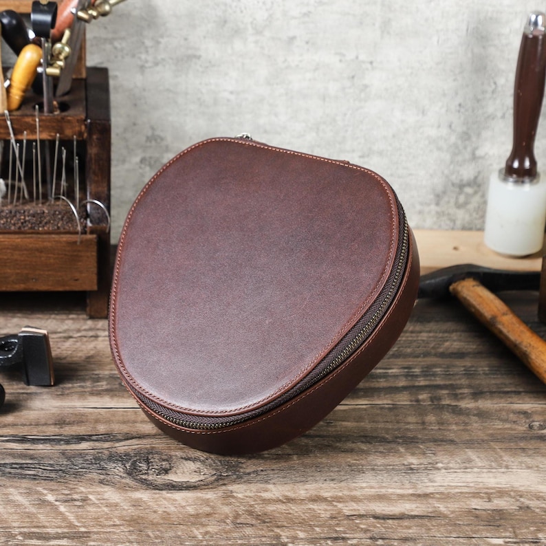 Op de afbeelding: Een bruin, ovaalvormig leren etui met een ritssluiting. De etui is gemaakt van een glad materiaal en ligt op een houten oppervlak. De etui is ontworpen voor het opbergen van kleine items.