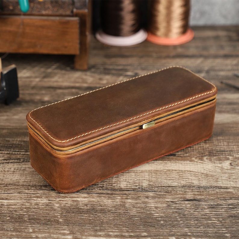 May include: A rectangular brown leather box with a hinged lid and a brass clasp. The box is stitched with white thread and sits on a wooden surface. The box appears to be a storage container.