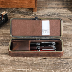 Peut inclure: &Eacute;tui &agrave; stylos en cuir marron ouvert avec fermoir en m&eacute;tal, contenant trois stylos. L'int&eacute;rieur de l'&eacute;tui est marron et doux, avec une petite poche pour une carte. Les stylos sont noirs, marron et rouges avec des d&eacute;tails argent&eacute;s.