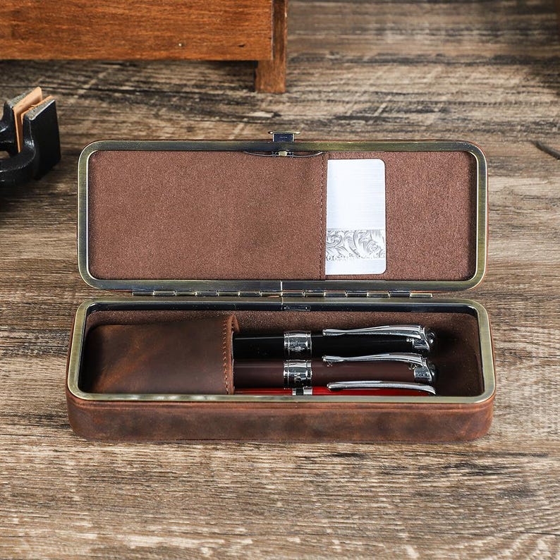 Peut inclure: &Eacute;tui &agrave; stylos en cuir marron ouvert avec fermoir en m&eacute;tal, contenant trois stylos. L'int&eacute;rieur de l'&eacute;tui est marron et doux, avec une petite poche pour une carte. Les stylos sont noirs, marron et rouges avec des d&eacute;tails argent&eacute;s.