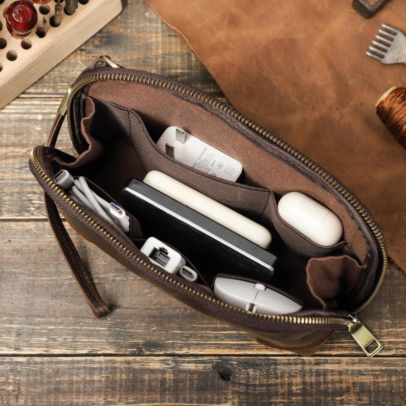 May include: A brown leather zippered pouch, open to reveal multiple compartments holding various tech accessories. Items include a white charger, a tablet, a white mouse, and a pair of wireless earbuds. The pouch is on a wooden surface.