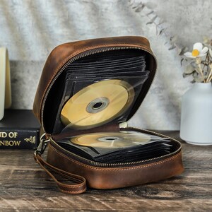 May include: A brown leather CD case, open to reveal several CDs in clear plastic sleeves. The case has a zipper closure and a small wrist strap. The case is sitting on a wooden surface.