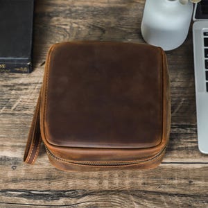 May include: A square, brown leather travel case with a zipper closure and a small handle. The case is sitting on a wooden surface. The leather has a distressed look.