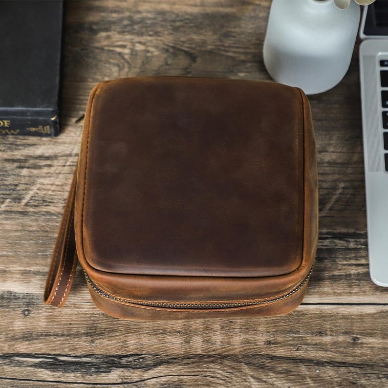 May include: A square, brown leather travel case with a zipper closure and a small handle. The case is sitting on a wooden surface. The leather has a distressed look.