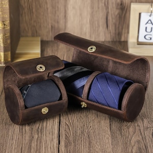 Può includere: Custodie per cravatte in pelle marrone con chiusure a scatto. Una custodia contiene una cravatta blu navy e un'altra una cravatta a righe blu. Le custodie sono aperte, rivelando le cravatte all'interno.