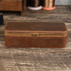 May include: A rectangular brown leather box with a hinged lid and a brass-colored clasp. The box is stitched with white thread and has a reddish-brown interior. The box is sitting on a wooden surface.