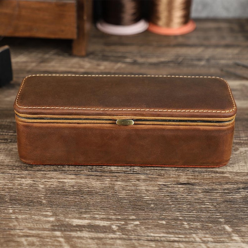 May include: A rectangular brown leather box with a hinged lid and a brass-colored clasp. The box is stitched with white thread and has a reddish-brown interior. The box is sitting on a wooden surface.