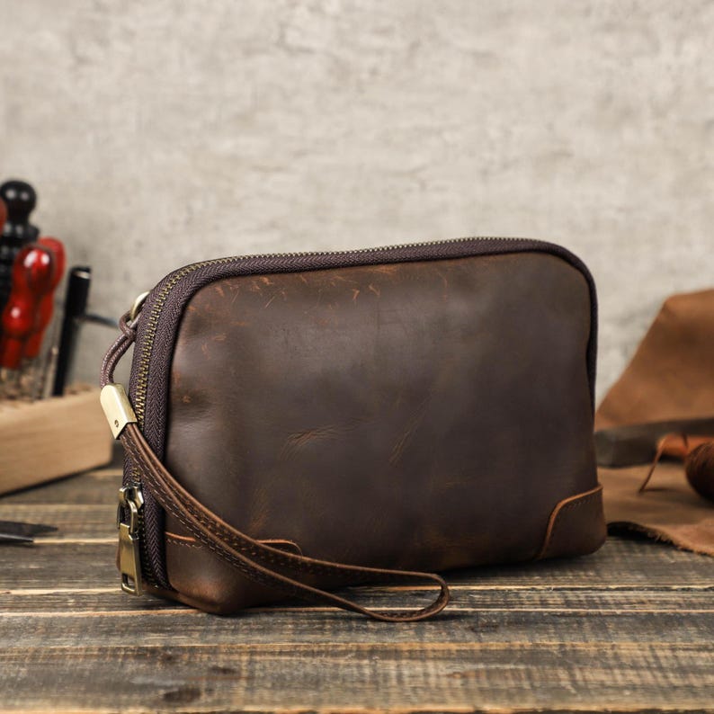 May include: A brown leather clutch with a zipper closure and a wrist strap. The rectangular bag has rounded corners and a dark brown zipper. The leather appears distressed, giving it a vintage look. The bag is sitting on a wooden surface.