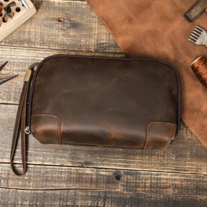 May include: A brown leather pouch with a wrist strap. The rectangular pouch has rounded corners and a zipper closure. The leather has a distressed appearance, with variations in color and texture. The pouch is on a wooden surface.