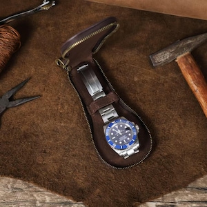 May include: A brown leather watch travel case with a silver and blue wristwatch inside. The case is open, revealing the watch secured by a strap. The watch has a blue bezel and a silver band. The case is on a brown surface.