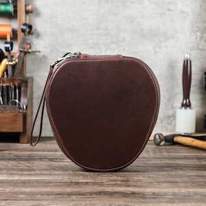 Op de afbeelding: Een bruine leren ovale etui met ritssluiting en polsbandje. Het ligt op een houten oppervlak. De achtergrond toont gereedschap, wat wijst op een leerambacht thema. Ongeveer 20 cm in diameter.