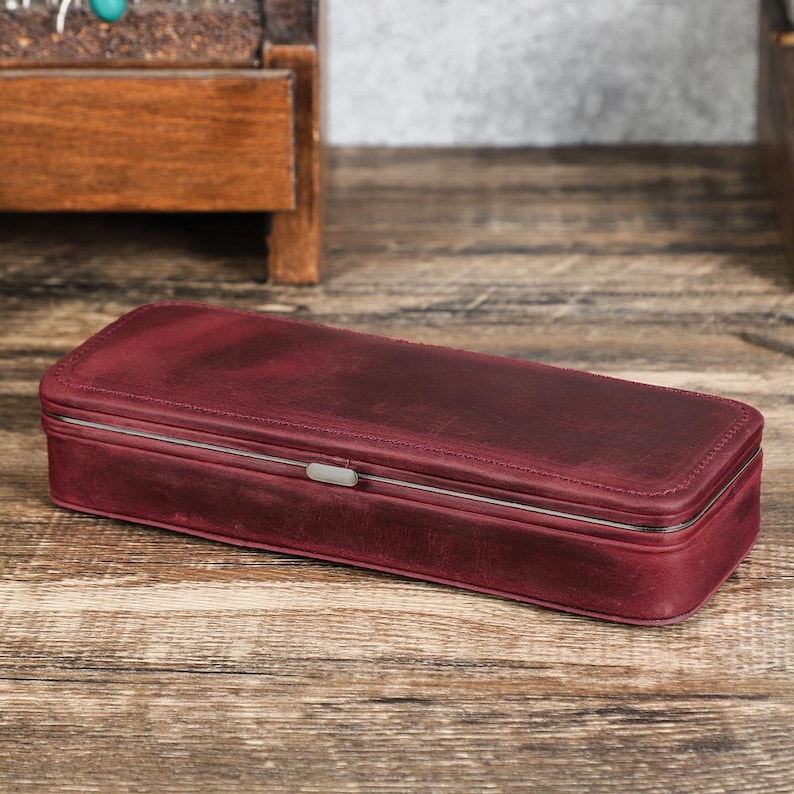 Peut inclure: Un &eacute;tui rectangulaire en cuir bordeaux avec un cadre en m&eacute;tal argent&eacute; et un petit fermoir. L'&eacute;tui est pos&eacute; sur une surface en bois. Le cuir a un aspect vieilli et les coutures sont visibles sur les bords. L'&eacute;tui est ferm&eacute;.