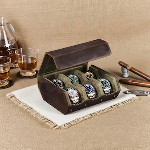 May include: A brown leather watch case with an open lid, revealing several wristwatches with various colored faces and bands. The case is lined with green fabric. Cigars and glasses of amber liquid are in the background.