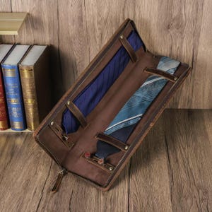 Può includere: Custodia per cravatte in pelle marrone aperta, con due cravatte. La custodia ha una chiusura a zip e cinturini in pelle. Una cravatta è blu a righe, l'altra è turchese con un motivo geometrico. Su superficie in legno.