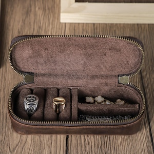 Może przedstawiać: Otwarty brązowy skórzany futerał podróżny na biżuterię z zamkiem błyskawicznym. Wewnątrz znajdują się przegródki na pierścionki i naszyjniki. Etui leży na drewnianej powierzchni.
