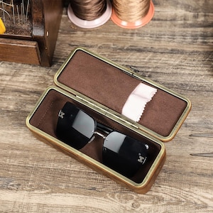May include: Open wooden eyeglasses case with a brown interior and a pair of black sunglasses. The case has a brass-colored metal frame and a small white cleaning cloth. The sunglasses have a silver frame.