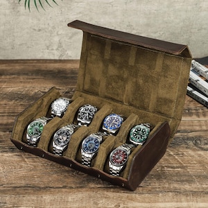 May include: A brown leather watch case with eight compartments, each holding a silver wristwatch with various colored watch faces. The case is open, revealing the watches against a green suede interior. The case is on a wooden surface.