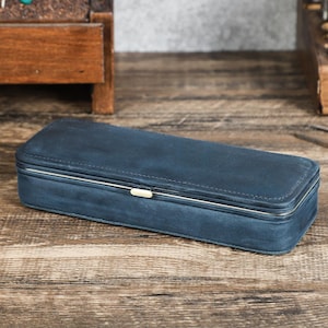 Peut inclure: Un &eacute;tui rectangulaire en cuir bleu marine avec une bordure en m&eacute;tal argent&eacute; et un petit fermoir dor&eacute;. L'&eacute;tui ferm&eacute; repose sur une surface en bois. Il semble &ecirc;tre destin&eacute; au rangement de petits objets.