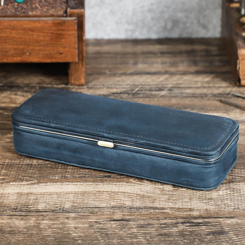 Peut inclure: Un &eacute;tui rectangulaire en cuir bleu marine avec une bordure en m&eacute;tal argent&eacute; et un petit fermoir dor&eacute;. L'&eacute;tui ferm&eacute; repose sur une surface en bois. Il semble &ecirc;tre destin&eacute; au rangement de petits objets.