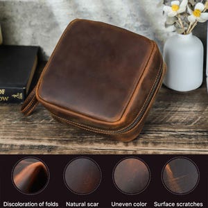 May include: A square, brown leather travel case with a wraparound zipper and a small handle. The leather has a distressed appearance with natural scars and surface scratches. The case is sitting on a wooden surface.