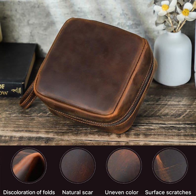 May include: A square, brown leather travel case with a wraparound zipper and a small handle. The leather has a distressed appearance with natural scars and surface scratches. The case is sitting on a wooden surface.