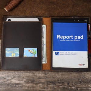 May include: A brown leather portfolio containing a notepad, credit cards, and cash. The notepad features a blue cover with the text "Report pad" and "Motivate higher office efficiency!". A pen holder is also visible.