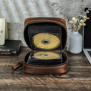 May include: Open brown leather CD case with a zipper closure, filled with several gold CDs in clear sleeves. The case is on a wooden surface, with a book and a laptop visible in the background.