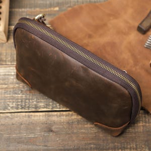May include: A brown leather pencil case with a brass zipper. The rectangular case has rounded corners and a small brass ring attached to the zipper. The leather has a distressed look.