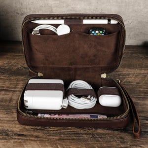 Könnte beinhalten: Ein offener, brauner Leder-Tech-Organizer, der verschiedene elektronische Zubehörteile zeigt. Darin befinden sich ein weißes Ladegerät, Kabel und weiße Ohrhörer. Der obere Bereich enthält einen Stift, Ladekabel und eine Smartwatch.