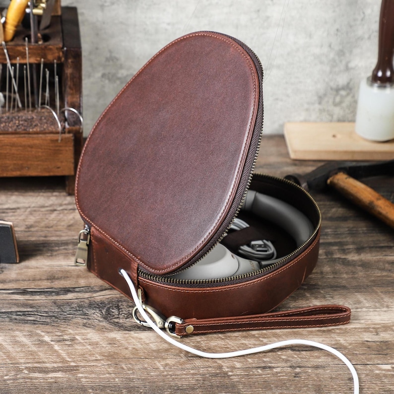 Op de afbeelding: Een bruine leren hoofdtelefoontas, open om een hoofdtelefoon en een kabel te laten zien. De tas heeft een ritssluiting, een polsband en een zilverkleurige sluiting. De achtergrond omvat gereedschap op een houten oppervlak.