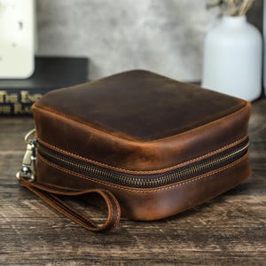 May include: A square, brown leather travel case with a zipper closure and a wrist strap. The case has a distressed finish, giving it a vintage look. The zipper is a dark bronze color.