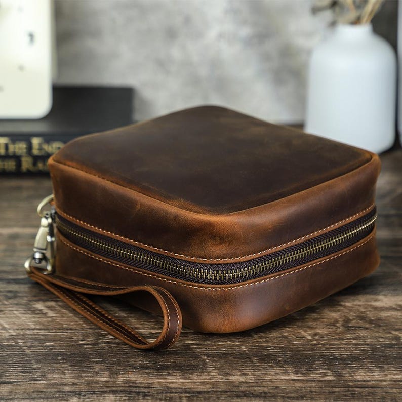 May include: A square, brown leather travel case with a zipper closure and a wrist strap. The case has a distressed finish, giving it a vintage look. The zipper is a dark bronze color.