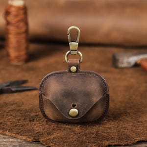 以下が含まれることがあります： 真鍮色の留め金とボタン留めが付いた、茶色の革製AirPodケース。ケースは使い古したような仕上がりで、小さなフラップが付いています。ワイヤレスイヤホンを保護し、持ち運ぶように設計されています。