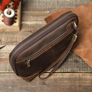May include: A brown leather clutch with a zipper closure and a wrist strap. The clutch has a rectangular shape and a textured surface. The zipper and hardware are gold-colored. The background includes leather and wooden elements.