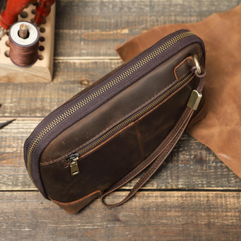 May include: A brown leather clutch with a zipper closure and a wrist strap. The clutch has a rectangular shape and a textured surface. The zipper and hardware are gold-colored. The background includes leather and wooden elements.
