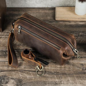 Op de afbeelding: Een bruine leren toilettas met twee compartimenten met ritssluiting en een polsband. Een tabakspijp ligt naast de tas in een lederen houder, bevestigd aan een sleutelhanger. De items liggen op een houten oppervlak.