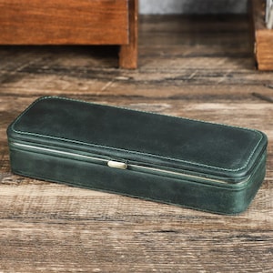 Peut inclure: Un &eacute;tui rectangulaire en cuir vert fonc&eacute; avec un fermoir et une garniture en m&eacute;tal argent&eacute;. L'&eacute;tui est ferm&eacute; et pr&eacute;sente des coutures blanches sur les bords. L'&eacute;tui est pos&eacute; sur une surface en bois.