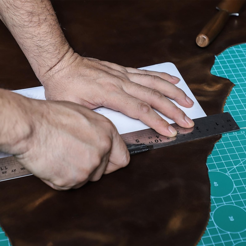 Op de afbeelding: Een persoon gebruikt een metalen liniaal om een patroon op bruin leer te meten en te snijden. Het leer ligt op een groene snijmat. Een houten gereedschap bevindt zich op de achtergrond.