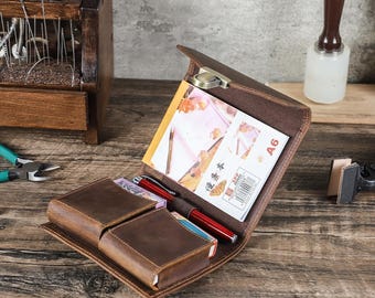 May include: Open brown leather wallet with a metal clasp, holding a notepad, two red pens, and two small boxes. The wallet is open on a wooden surface, with a wooden tool stand in the background.