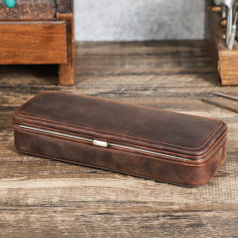 Peut inclure: Un &eacute;tui rectangulaire en cuir marron avec un cadre en m&eacute;tal argent&eacute; et un fermoir. L'&eacute;tui est ferm&eacute; et repose sur une surface en bois. Le cuir a un aspect vieilli avec des coutures visibles.