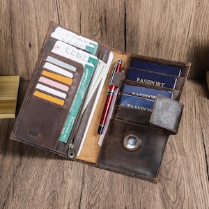 May include: Open brown leather travel wallet with multiple compartments. The wallet contains credit cards, boarding passes, cash, and passports. A red pen rests inside the wallet. The wallet has an RFID tag.
