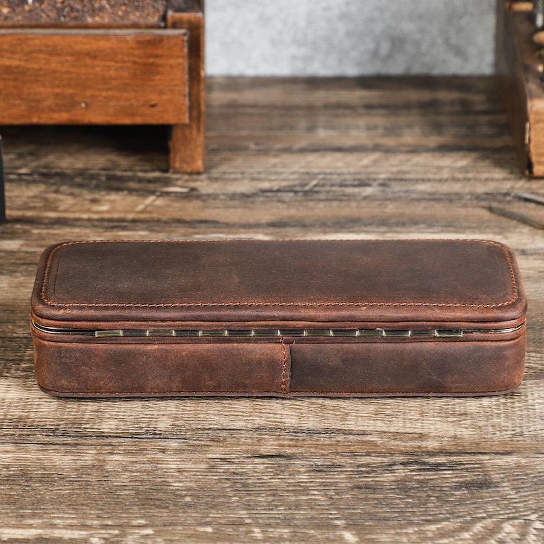 Peut inclure: Un &eacute;tui rectangulaire en cuir marron avec un couvercle &agrave; charni&egrave;re. L'&eacute;tui est cousu sur les bords et poss&egrave;de une charni&egrave;re m&eacute;tallique. Le cuir a un aspect vieilli. L'&eacute;tui est pos&eacute; sur une surface en bois.