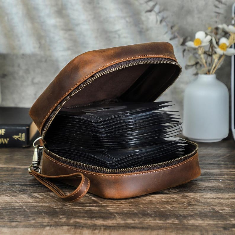 May include: A brown leather CD case with a zipper closure, open to reveal a stack of black CD sleeves. The case has a handle and a silver-colored clasp. The background includes a vase with white flowers.