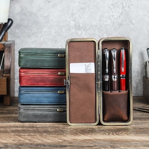 Peut inclure: Plusieurs &eacute;tuis &agrave; stylos en cuir de diff&eacute;rentes couleurs : vert, rouge, bleu et gris. Un &eacute;tui est ouvert, r&eacute;v&eacute;lant trois stylos et une carte. L'&eacute;tui ouvert a un int&eacute;rieur marron avec une poche pour cartes et des porte-stylos. Les stylos sont noirs et argent&eacute;s, et rouge.