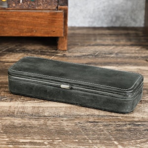 Peut inclure: Un &eacute;tui rectangulaire en daim gris fonc&eacute; avec des coutures blanches et un fermoir argent&eacute;. L'&eacute;tui est ferm&eacute; et repose sur une surface en bois. L'&eacute;tui semble &ecirc;tre destin&eacute; au rangement d'objets.