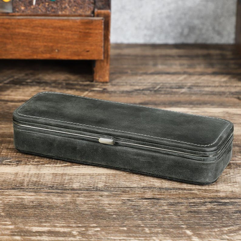 Peut inclure: Un &eacute;tui rectangulaire en daim gris fonc&eacute; avec des coutures blanches et un fermoir argent&eacute;. L'&eacute;tui est ferm&eacute; et repose sur une surface en bois. L'&eacute;tui semble &ecirc;tre destin&eacute; au rangement d'objets.