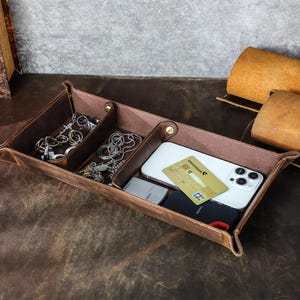 Könnte beinhalten: Ein braunes Leder-Valet-Tablett mit drei Fächern. Das Tablett enthält Schmuck, ein Smartphone, eine Kreditkarte und ein kleines Feuerzeug. Das Tablett dient zum Organisieren und Aufbewahren von Alltagsgegenständen.