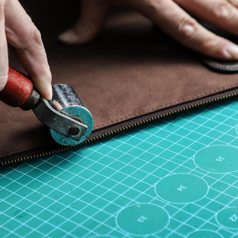 Op de afbeelding: Een close-up van een leerambachtelijk project in uitvoering. Een persoon gebruikt een rolwerktuig met een rode handgreep om de rits van een bruin lederen item aan te drukken. Het werkoppervlak is een groene snijmat.
